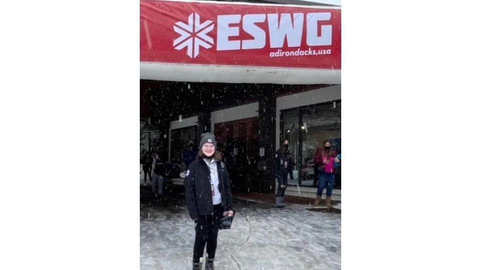 Girl wearing a black coat standing under a red sign with the ESWG logo on it.